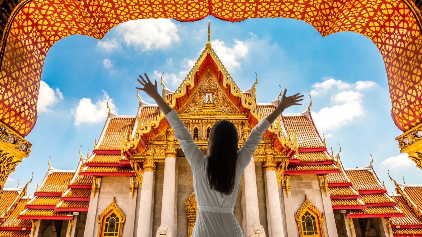 A traveller stands with both arms raised in front of the gilded roofs and white columns of the Marble Temple in Bangkok